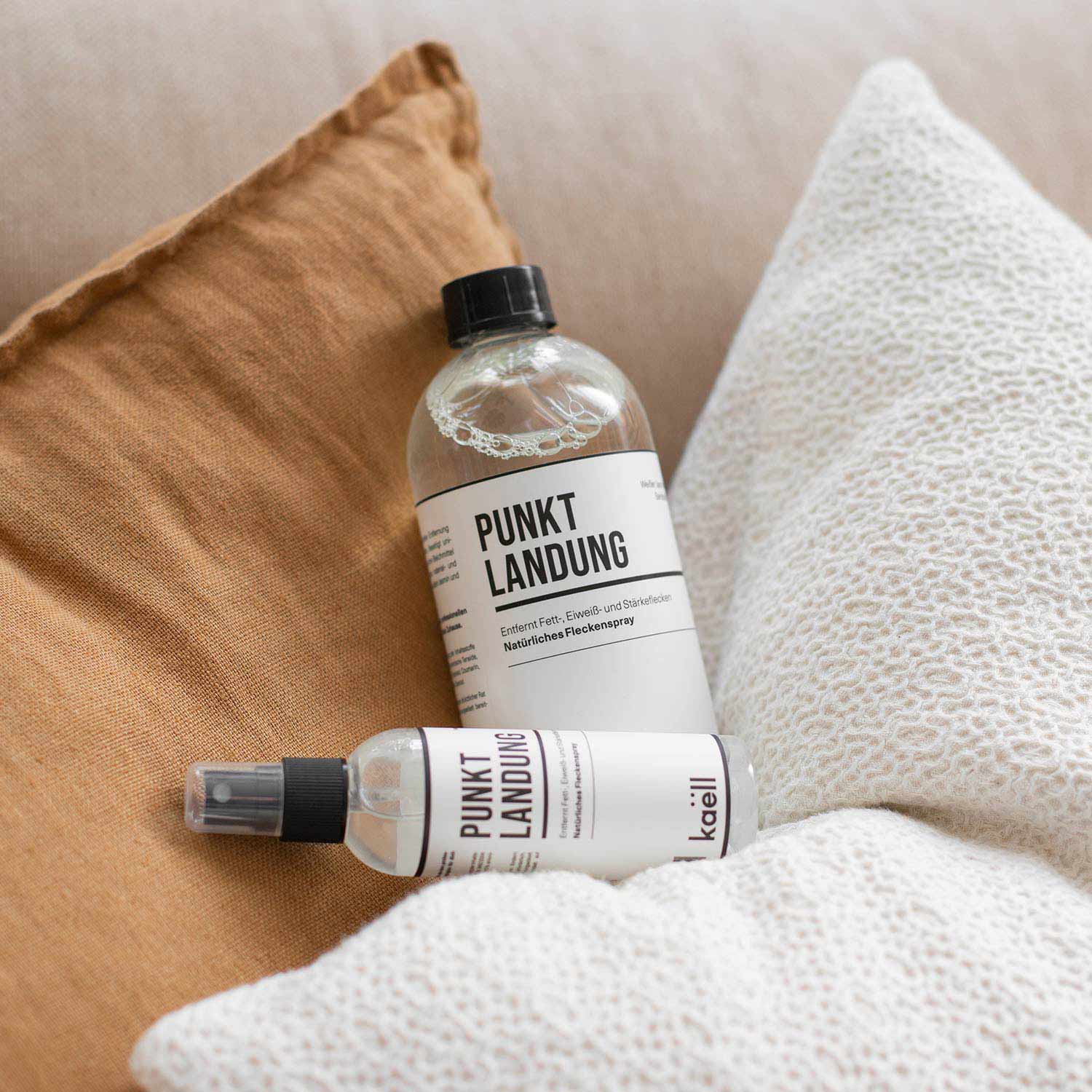 Zwei Produkte mit der Aufschrift 'Punktlandung', bestehend aus einer 500ml Flasche und einem 100ml Spray, liegen dekorativ auf einem braunen Kissen. Im Hintergrund ist ein weiß strukturiertes Kissen zu sehen. Oben links im Bild befindet sich ein orangefarbenes Feld mit dem Schriftzug 'NEU'.