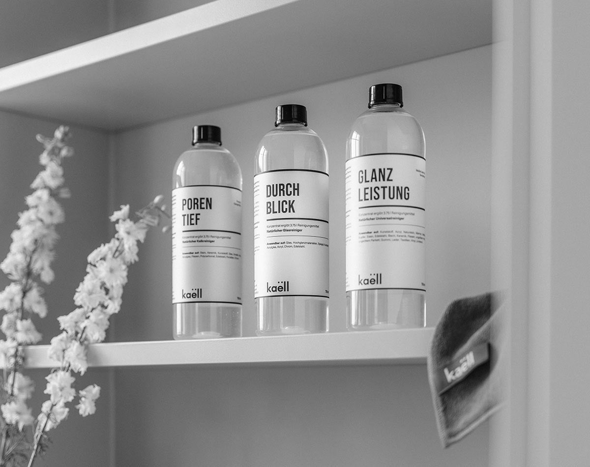 Drei Flaschen Reinigungsmittel mit den Aufschriften 'Porentief', 'Durchblick' und 'Glanzleistung' stehen ordentlich nebeneinander auf einem Regal. Links im Bild befinden sich dekorative Zweige mit Blüten, und rechts ist ein zusammengefaltetes Reinigungstuch mit 'kaëll'-Label zu sehen.