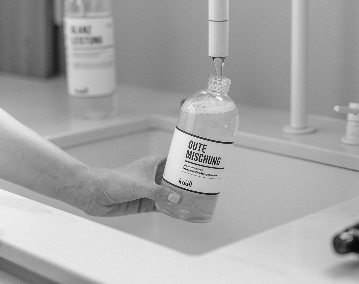 Eine Hand hält eine Flasche mit der Aufschrift 'Gute Mischung' unter einen Wasserhahn, während die Flasche mit Wasser befüllt wird. Im Hintergrund ist eine weitere Flasche mit der Aufschrift 'Glanzleistung' unscharf zu erkennen. Die Szene spielt sich in einer Küchenspüle ab.