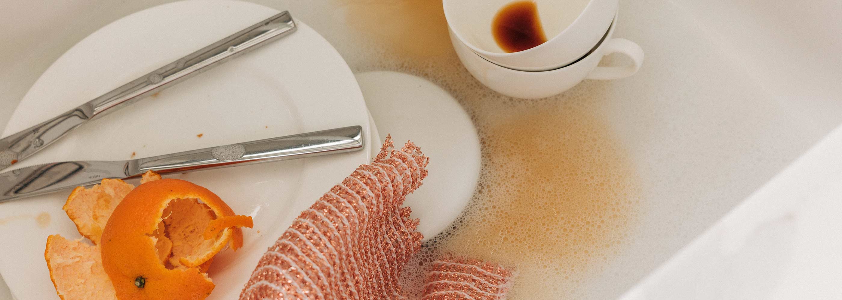 Ein Spülbecken mit schmutzigem Geschirr: ein Teller mit Messer und Gabel, Orangenschalen, eine weiße Tasse mit Kaffeeresten und ein nasses Kupfertuch in einem Wasserbad mit Seifenblasen. Hier findest du die Schritt für Schritt Anleitungen für die Reinigung von: Backöfen, Dunstabzugshauben, Kühlschranken, Wasserkochern, Spülmaschinen und Küchenschränken.